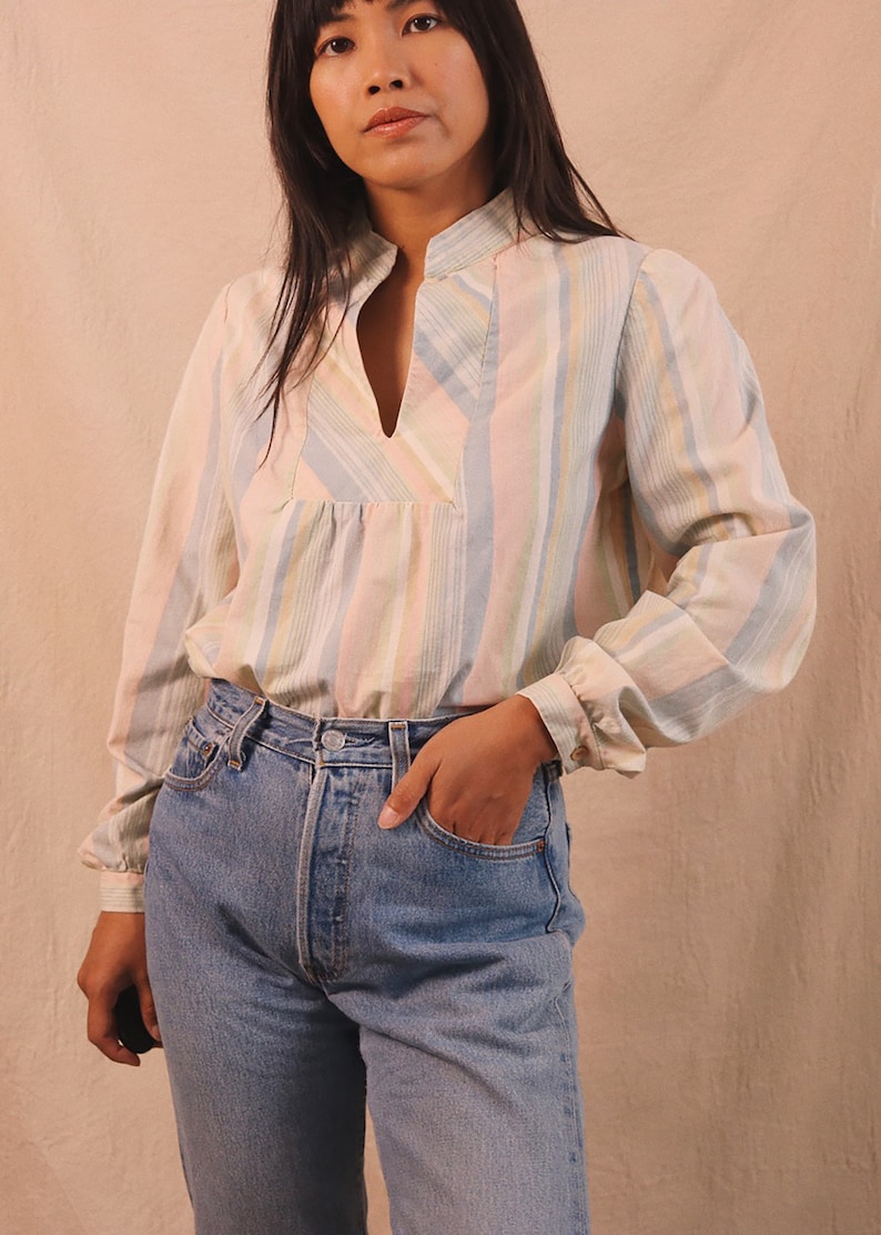 Peut inclure: Un chemisier &agrave; rayures pastel avec des manches longues et un d&eacute;collet&eacute; en V. Le chemisier pr&eacute;sente des rayures verticales dans les tons de rose, bleu, jaune et blanc. Le mod&egrave;le porte un jean en denim bleu.