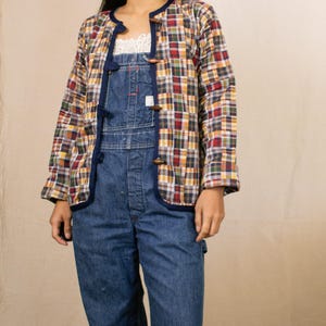 May include: A colorful plaid jacket with a navy blue trim and wooden toggle closures. The jacket is worn over blue denim overalls. The plaid pattern includes red, yellow, green, and white squares. The outfit is casual and vintage-inspired.