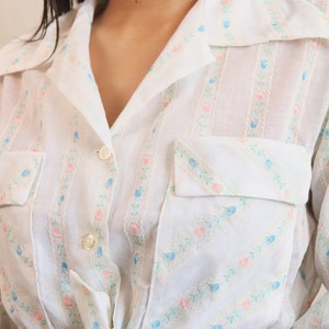 May include: White button-up shirt with a floral pattern in pink and blue. The shirt has a collared neckline, two chest pockets, and is tied at the waist. The floral design is arranged in vertical stripes.