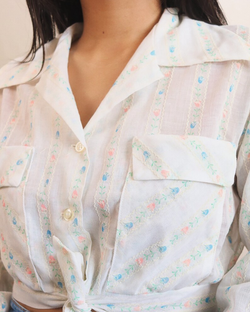 May include: White button-up shirt with a floral pattern in pink and blue. The shirt has a collared neckline, two chest pockets, and is tied at the waist. The floral design is arranged in vertical stripes.