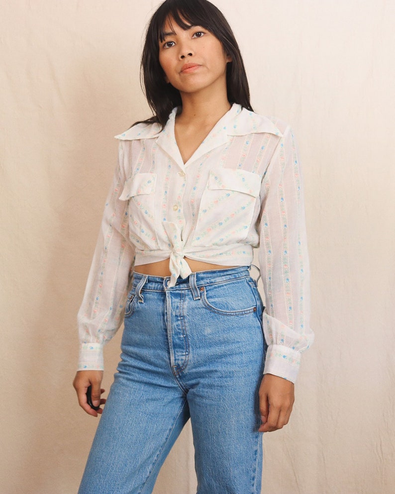May include: A white, long-sleeved shirt with a tie-front design. The shirt features a delicate floral pattern in pink and blue, and two front pockets. Paired with light blue denim jeans.