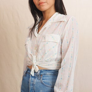 May include: A white button-down shirt with a floral pattern in pink, blue, and green. The shirt has two front pockets and is tied at the waist. The model is wearing blue denim jeans.