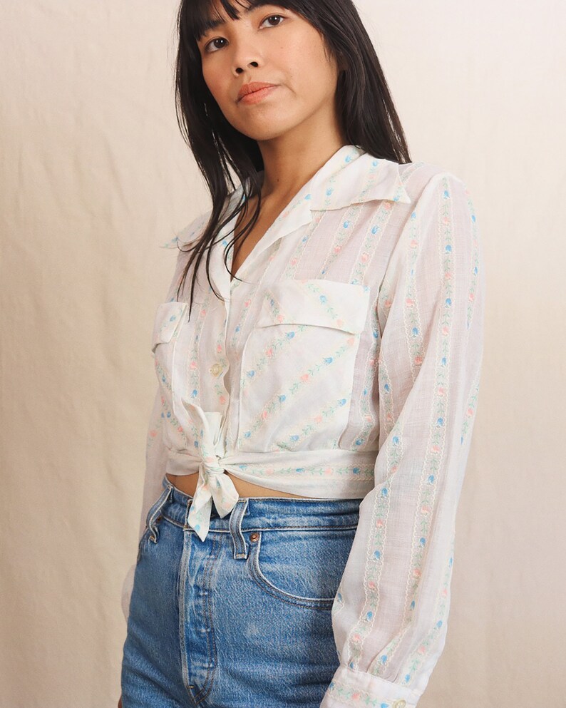 May include: A white button-down shirt with a floral pattern in pink, blue, and green. The shirt has two front pockets and is tied at the waist. The model is wearing blue denim jeans.