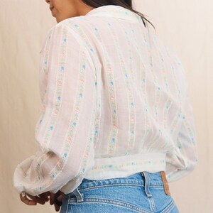 May include: White blouse with a floral pattern in pink, blue, and green. The blouse has long sleeves and a collared neckline. The model is wearing blue jeans.