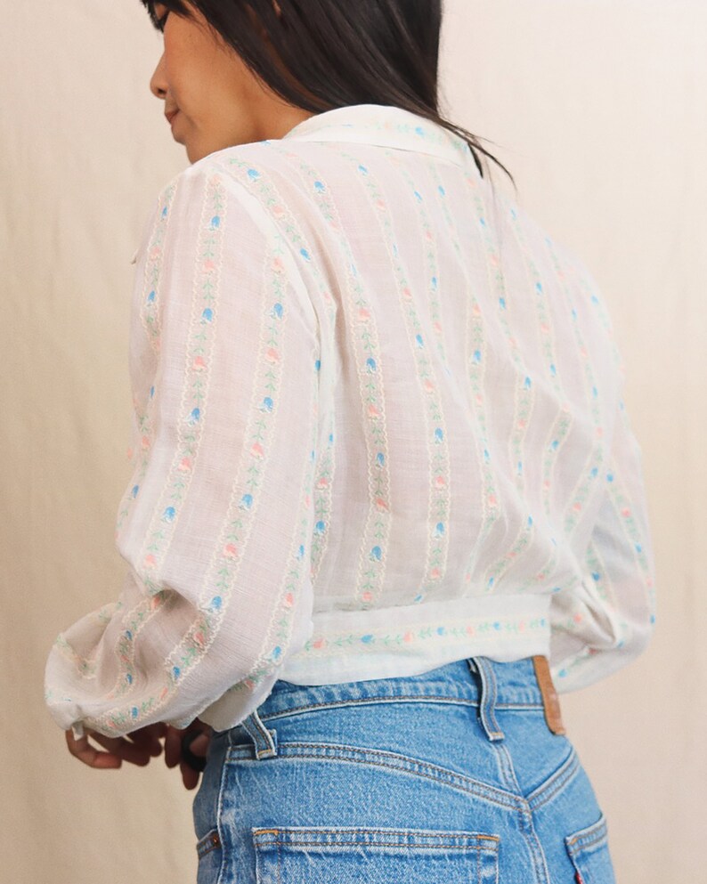 May include: White blouse with a floral pattern in pink, blue, and green. The blouse has long sleeves and a collared neckline. The model is wearing blue jeans.