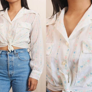 May include: White button-up shirt with long sleeves, two chest pockets, and a tie-front closure. The shirt features a vertical striped pattern with small pink and blue floral accents. The collar is a classic style.