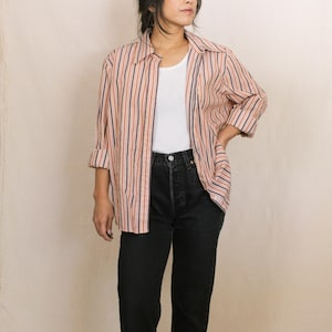 May include: A button-down shirt with vertical stripes in shades of orange, brown, and navy blue. The shirt is worn over a white tank top and paired with black denim jeans. The sleeves are rolled up, and the outfit is casual.