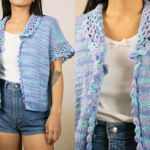 Puede incluir: Cárdigan de manga corta en tonos azul pastel y lila. El tejido de punto abierto presenta un cuello y bordes de manga festoneados, con un detalle calado. El cárdigan se combina con pantalones cortos vaqueros.