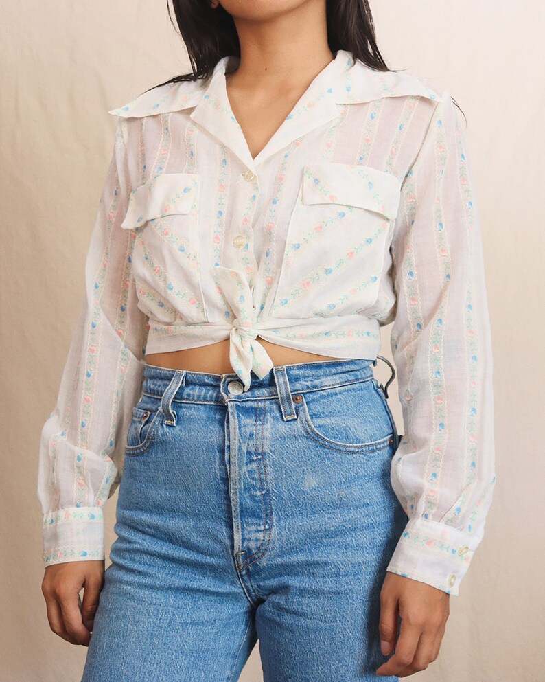 May include: White button-up shirt with a floral pattern and two chest pockets, tied at the waist. The shirt is paired with light blue denim jeans. The shirt features a delicate pattern of pink and blue flowers.