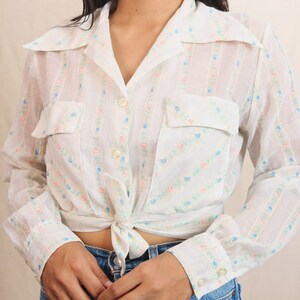 May include: White button-up shirt with a floral pattern in pink and blue. The shirt has two front pockets, long sleeves, and a tie at the waist. The shirt is paired with blue jeans.