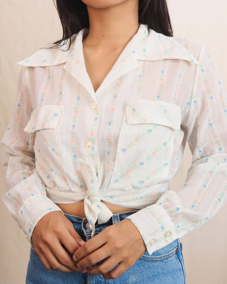 May include: White button-up shirt with a floral pattern in pink and blue. The shirt has two front pockets, long sleeves, and a tie at the waist. The shirt is paired with blue jeans.