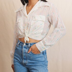 May include: A white, long-sleeved blouse with a floral pattern and two front pockets. The blouse is tied at the waist and paired with light blue denim jeans. The floral pattern includes pink and blue accents.