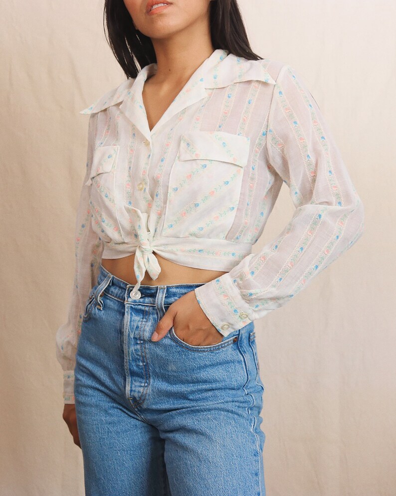 May include: A white, long-sleeved blouse with a floral pattern and two front pockets. The blouse is tied at the waist and paired with light blue denim jeans. The floral pattern includes pink and blue accents.