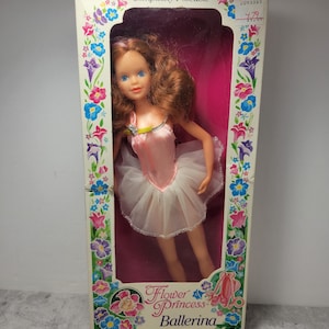 May include: A vintage Flower Princess Ballerina doll in a pink and white tutu, with brown hair and blue eyes. The doll is posed in a ballet position and is packaged in a box with a floral design. The box also includes the text "Completely Poseable", "Quality made 11 1/2" tall", and "creata".