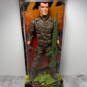 May include: An Ultra Elite action figure in a clear plastic package. The figure is dressed in a brown military uniform and holds a green rifle. The packaging reads "THE CORPS! ULTRA ELITE SOLO PURSUIT! 26.5CM FIGURE!" and "TRACKER FIGURAS!"