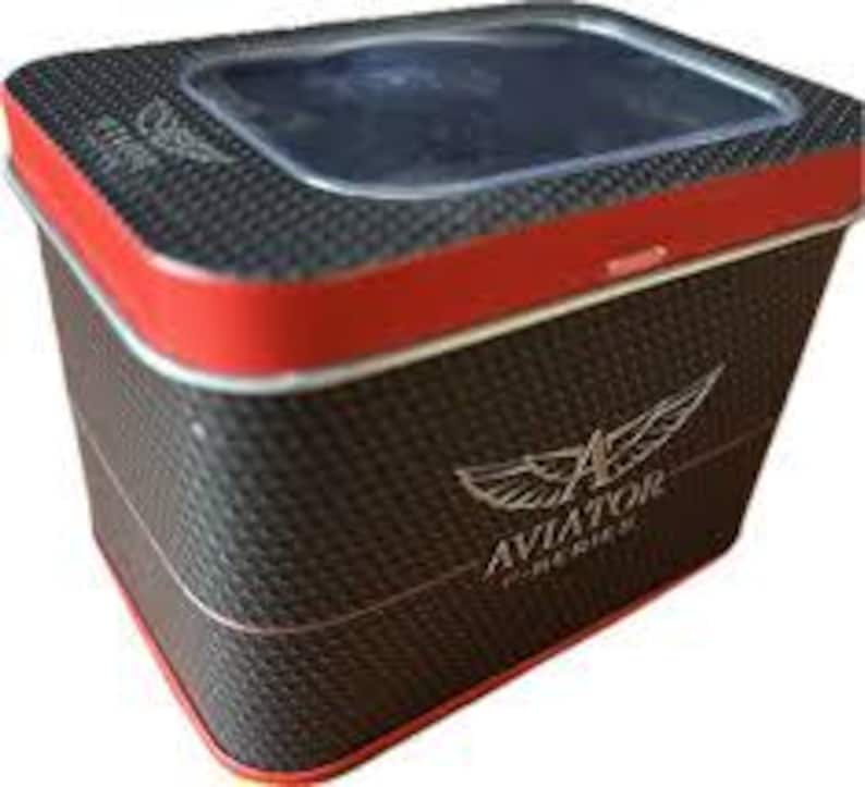 May include: A rectangular watch box with a carbon fibre pattern and red trim. The box has a clear top and the word "AVIATOR" with a wing logo is printed on the side.