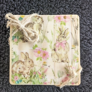 May include: A wooden sign with a floral pattern and bunnies. The sign is decorated with a brown twine bow.