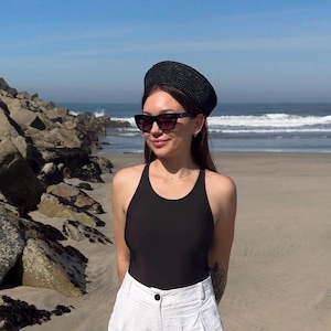 Puede incluir: Una boina negra texturizada, gafas de sol negras con cristales oscuros, un body negro sin mangas con cuello redondo y pantalones cortos blancos de talle alto con un botón visible y pliegues frontales.