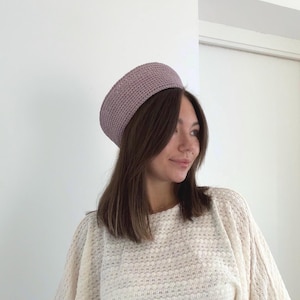 Puede incluir: Un sombrero de color malva en forma de cúpula, elaborado con un patrón de punto de ganchillo denso y uniforme. El sombrero presenta una corona redondeada y estructurada con un borde circular limpio que se ajusta firmemente, mostrando una textura de hilo consistente.