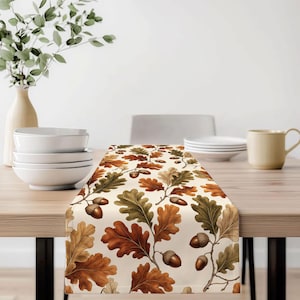 Può includere: Un runner da tavola rettangolare con un motivo ripetuto di foglie di quercia marroni, beige e verdi intervallate da ghiande marroni su uno sfondo color crema. Il design mostra dettagliate illustrazioni botaniche di fogliame e frutti.