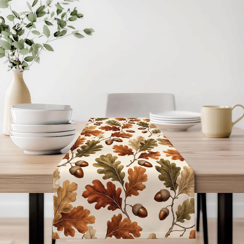 May include: A rectangular table runner featuring a repeating pattern of brown, tan, and green oak leaves interspersed with brown acorns on a cream-colored background. The design displays detailed botanical illustrations of foliage and nuts.