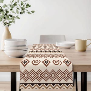 Könnte beinhalten: Beiger Tischläufer mit einem sich wiederholenden geometrischen Muster in Brauntönen. Das Design umfasst Rauten, Zickzackmuster und Spiralformen. Der Läufer wird auf einem Holztisch präsentiert, mit einer Vase mit Grün und Geschirr im Hintergrund.