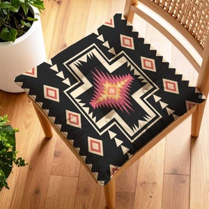 Op de afbeelding: Een vierkant stoelkussen met een zwarte achtergrond en een geometrisch patroon in crème, roze en rood. Het kussen ligt op een houten stoel met een geweven rugleuning, op een houten vloer.
