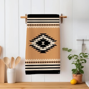 May include: A tan and black geometric patterned kitchen towel hanging on a wooden towel rack. The towel features a central diamond design and horizontal stripes. The towel is displayed in a kitchen setting with wooden utensils and a potted plant.