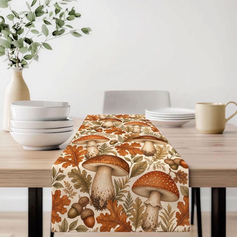 May include: A rectangular table runner featuring a repeating pattern of various mushrooms, brown oak leaves, and acorns in shades of orange, brown, and cream. The design includes detailed illustrations of fungi with spotted caps and stems.