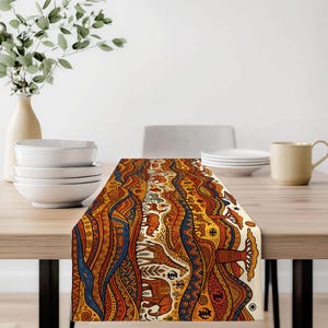 May include: A table runner with an African-inspired design. The runner features a repeating pattern of stylized animals, trees, and geometric shapes in shades of orange, brown, blue, and cream. It is placed on a wooden table.