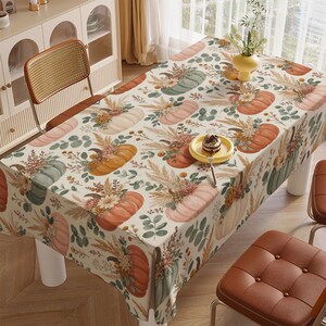 May include: A rectangular tablecloth featuring a repeating pattern of pumpkins in shades of orange, sage green, and cream, accented with dried floral arrangements, wheat stalks, and eucalyptus leaves on a light cream background.