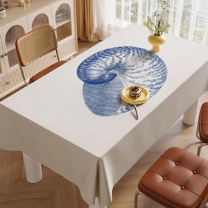 Può includere: Una tovaglia rettangolare bianca con un grande disegno di nautilus blu. Il tavolo è apparecchiato con un piattino giallo con un dolce. Sedie in pelle marrone e un mobile sullo sfondo.