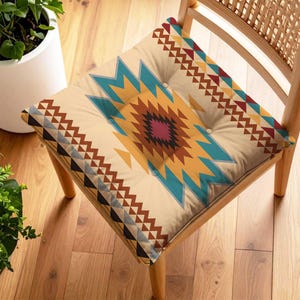 Pode incluir: Uma almofada de cadeira bege com um padrão geométrico em tons de azul-petróleo, castanho, amarelo e bordô. A almofada está numa cadeira de madeira com encosto entrançado. A almofada tem um design com botões.