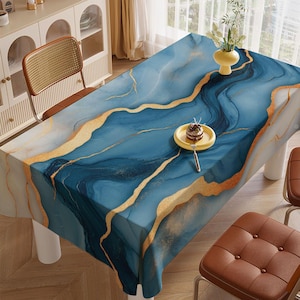 May include: A rectangular tablecloth with a blue and gold abstract design. The tablecloth features a marbled effect with flowing blue hues and gold accents. A small yellow plate with a dessert and a fork sits on the tablecloth. The table is surrounded by chairs.