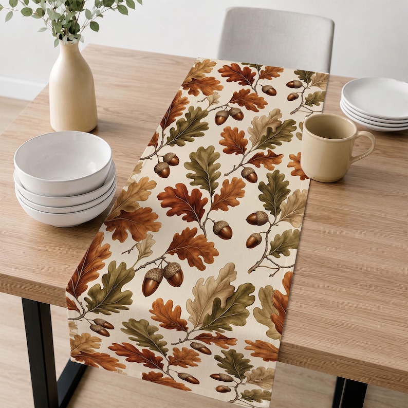 May include: A rectangular table runner featuring a repeating pattern of brown acorns and oak leaves in shades of green, tan, and orange on a cream background. The runner is laid flat across a wooden table surface with a stack of bowls and a mug.
