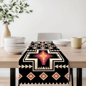 May include: A black table runner with a geometric pattern in cream, red, and yellow. The design features diamond and arrow shapes, with a central cross-like motif. The runner is displayed on a wooden table, showcasing its decorative appeal.