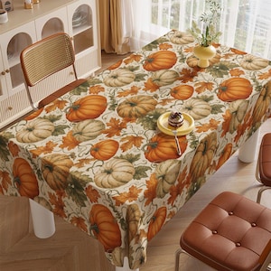 May include: A rectangular tablecloth featuring a repeating pattern of orange and pale green pumpkins, brown and orange autumn leaves, and winding vines on a cream background. A small yellow plate with a dessert sits on the table surface.