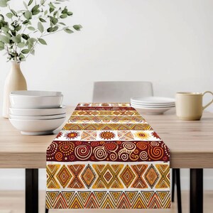 Puede incluir: Un camino de mesa decorativo con un patrón repetido de formas geométricas y diseños tribales en tonos naranja, amarillo y marrón. El camino de mesa está colocado sobre una mesa de madera, con platos blancos y un jarrón de vegetación en el fondo.