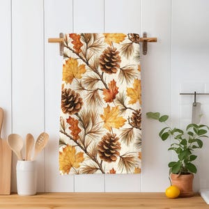 May include: A rectangular kitchen towel featuring a repeating pattern of brown pinecones, thin brown pine needles, and autumn maple leaves in shades of yellow, orange, and brown against a cream-colored background, hanging on a wooden rod.