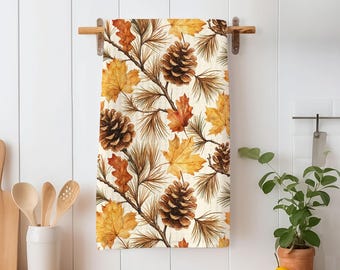 May include: A rectangular kitchen towel featuring a repeating pattern of brown pinecones, thin brown pine needles, and autumn maple leaves in shades of yellow, orange, and brown against a cream-colored background, hanging on a wooden rod.