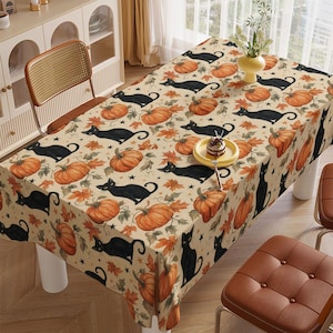 May include: A rectangular tablecloth featuring a repeating pattern of black cats, orange pumpkins, and autumn leaves on a light beige background. Small black stars are scattered throughout the design, and a small plate with a dessert sits on top.