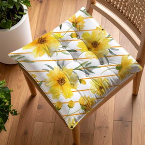 May include: A square chair cushion with a floral pattern. The cushion features large yellow flowers with green leaves and orange stripes on a white background. The cushion is button-tufted and sits on a wooden chair.