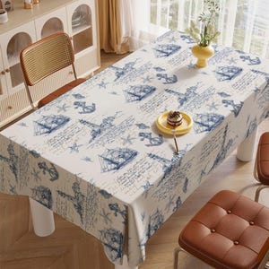May include: A rectangular tablecloth with a nautical design. The white cloth features blue illustrations of sailboats, anchors, and script. A yellow plate with a small cake sits on the table. The table has white legs and brown chairs.