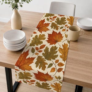 May include: A rectangular table runner featuring a repeating pattern of autumn leaves in shades of orange, red, yellow, and olive green on a cream background. The fabric runner is laid flat across a wooden table surface with a smooth texture.
