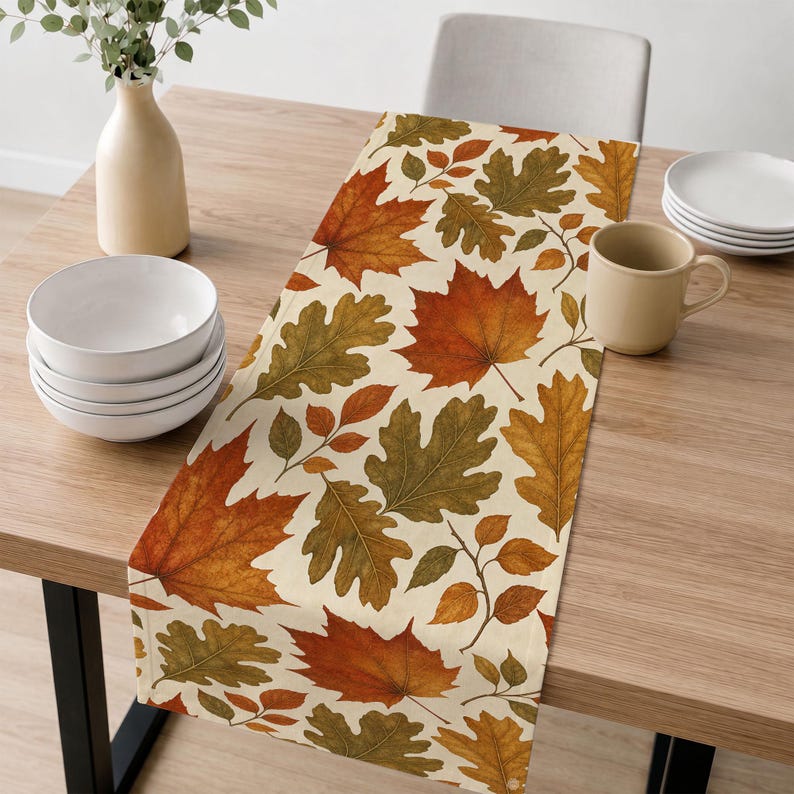 May include: A rectangular table runner featuring a repeating pattern of autumn leaves in shades of orange, red, yellow, and olive green on a cream background. The fabric runner is laid flat across a wooden table surface with a smooth texture.