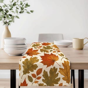 May include: A rectangular table runner featuring a repeating pattern of autumn leaves in shades of orange, yellow, brown, and olive green on a cream-colored background, draped across a wooden table surface with white bowls and a mug.