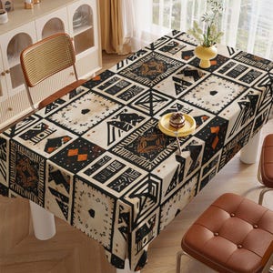 May include: Rectangular tablecloth with a geometric pattern in black, orange, and cream. The design features squares and rectangles with tribal-inspired motifs. A yellow plate with a small cake sits on the table, along with a vase of flowers.