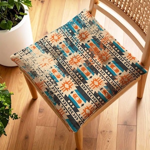 May include: Square chair cushion with a Southwestern-inspired design. The cushion features a beige background with a distressed texture and a pattern of geometric shapes in teal, orange, and black. The cushion is placed on a wooden chair.