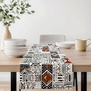 May include: A rectangular table runner with a repeating geometric pattern in black, brown, and cream. The design features various shapes and symbols, creating a tribal or ethnic aesthetic. The runner is placed on a wooden table.