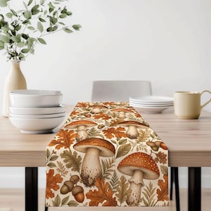 May include: A rectangular table runner featuring a repeating pattern of various mushrooms, brown oak leaves, and acorns in shades of orange, brown, and cream. The design includes detailed illustrations of fungi with spotted caps and stems.